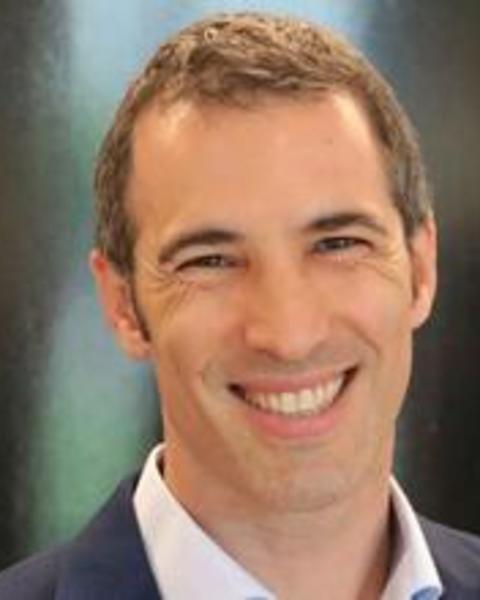 Headshot of Yossi Harpaz standing and smiling in front of a blurred background